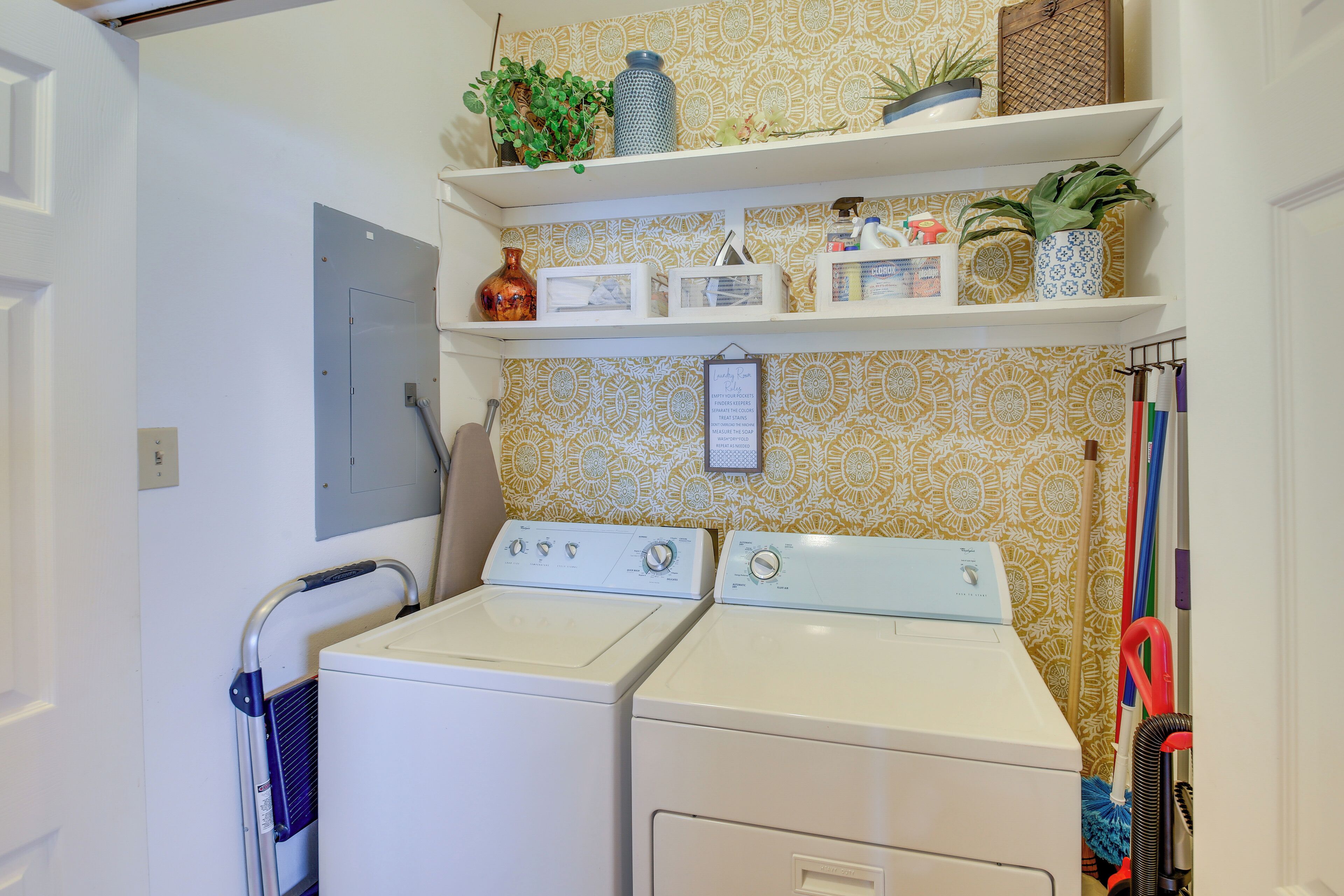 Une buanderie équipée de deux machines à laver et deux sèche-linge superposés, d’étagères ornées de plantes et d’objets décoratifs, et d’une porte sur laquelle est fixé un porte-serviettes.