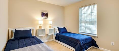 Une chambre avec deux lits, une fenêtre avec des stores, un luminaire fixé au mur et une petite étagère avec une lampe.