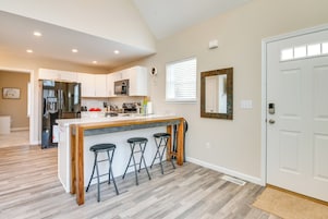 A modern kitchen with white cabinets, a stainless steel refrigerator, a wooden island with bar stools, and a large window with blinds.