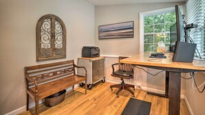 A home office with a wooden desk, a computer, a printer, a bench, and a decorative window with blinds.