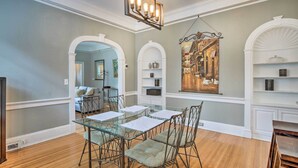 A dining area with a glass table and metal chairs, white shelving with decorative items, and a large framed painting of a street scene.