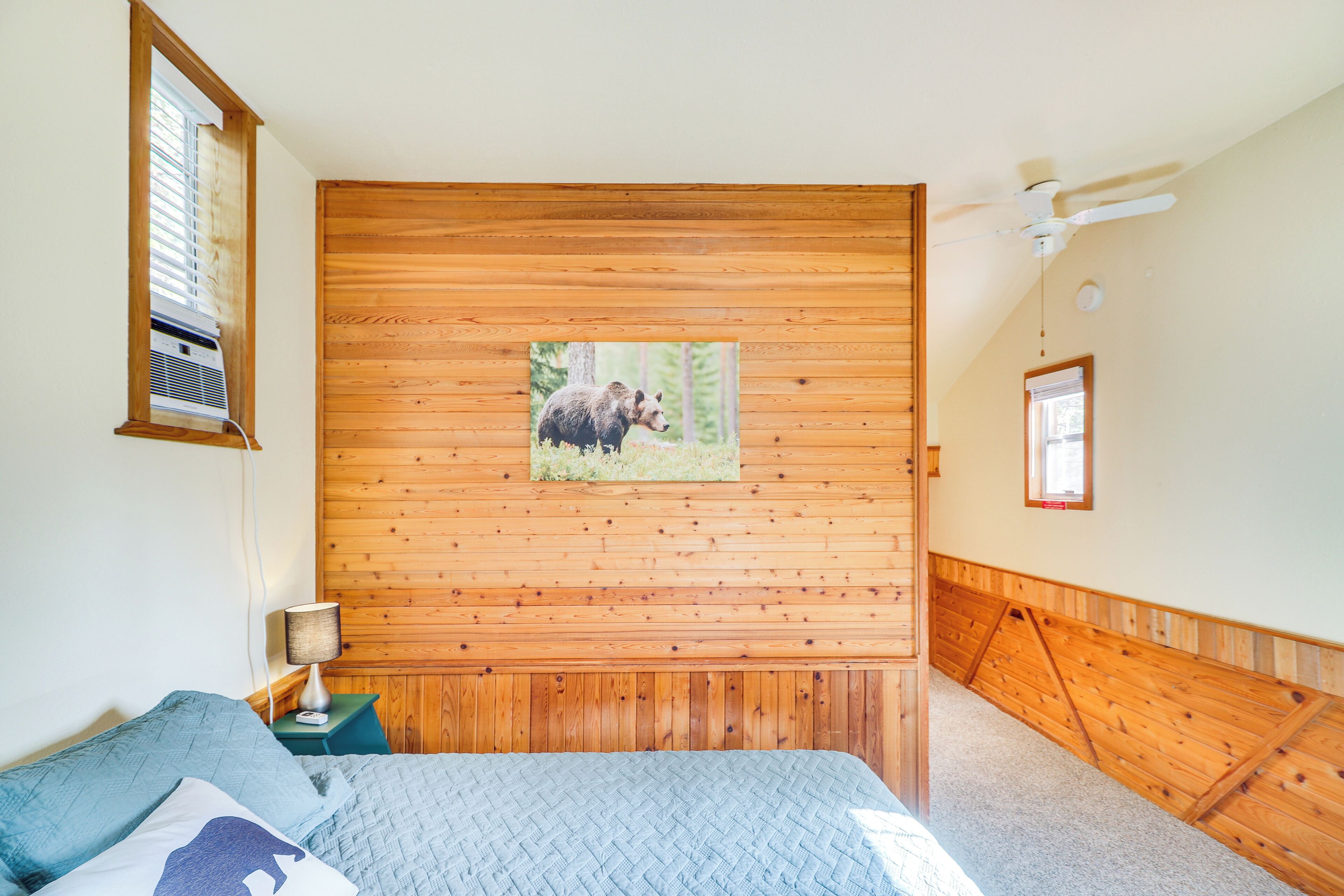 Ein Schlafzimmer mit Holz-Kopfteil, einem Bett mit blauer Bettwäsche, einem Nachttisch mit Lampe und einer Wand-Klimaanlage.