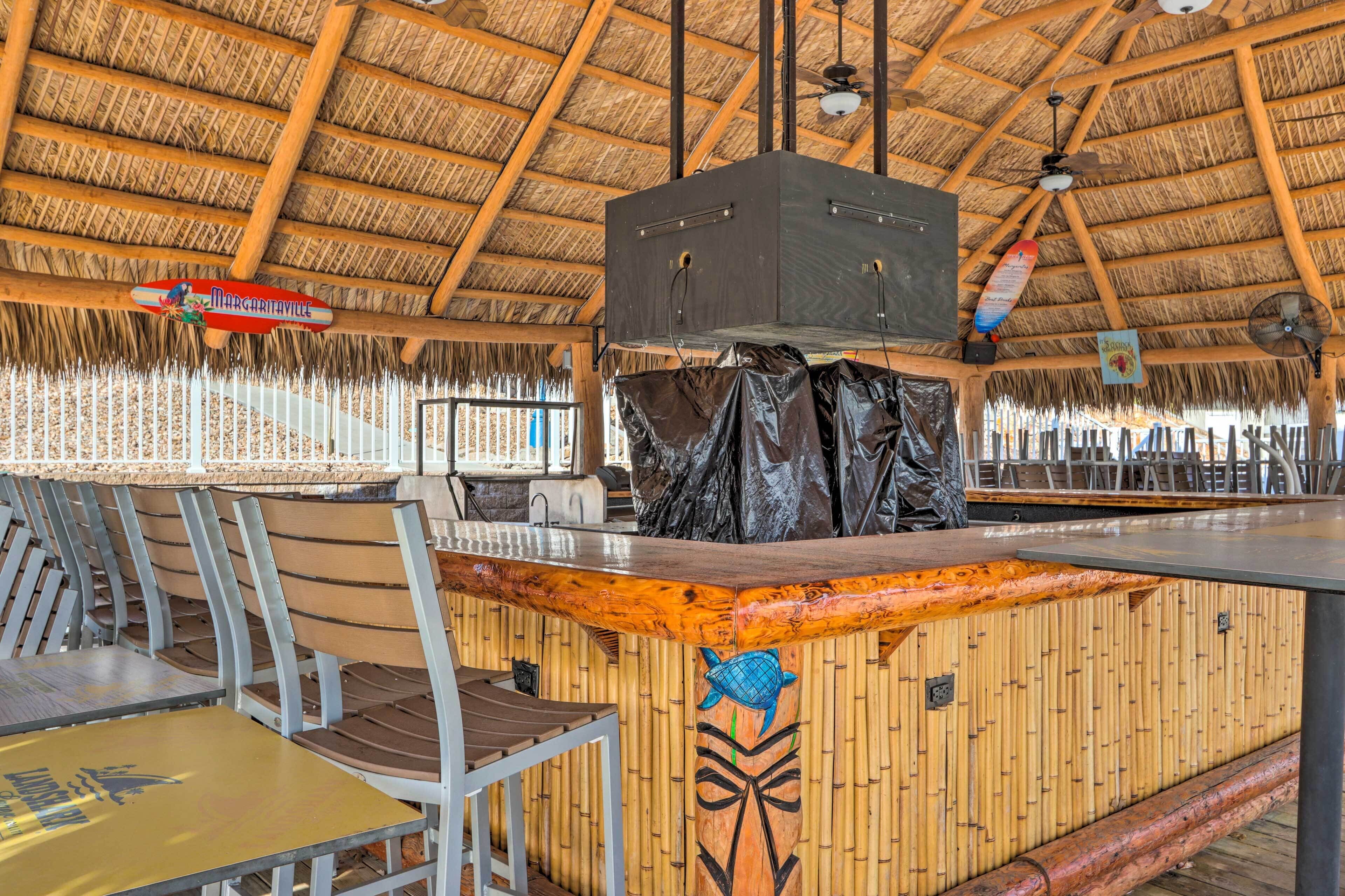 Um restaurante com telhado de palha, mesas e cadeiras de madeira, área de bar central e um mural decorativo com peixes.