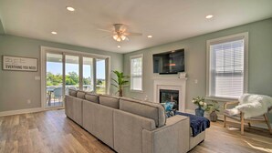 A spacious living room with a large gray sectional sofa, a flat-screen TV mounted above a fireplace, and a ceiling fan. - Gorgeous Newly Built Home: Golf Course View! (Brigantine)