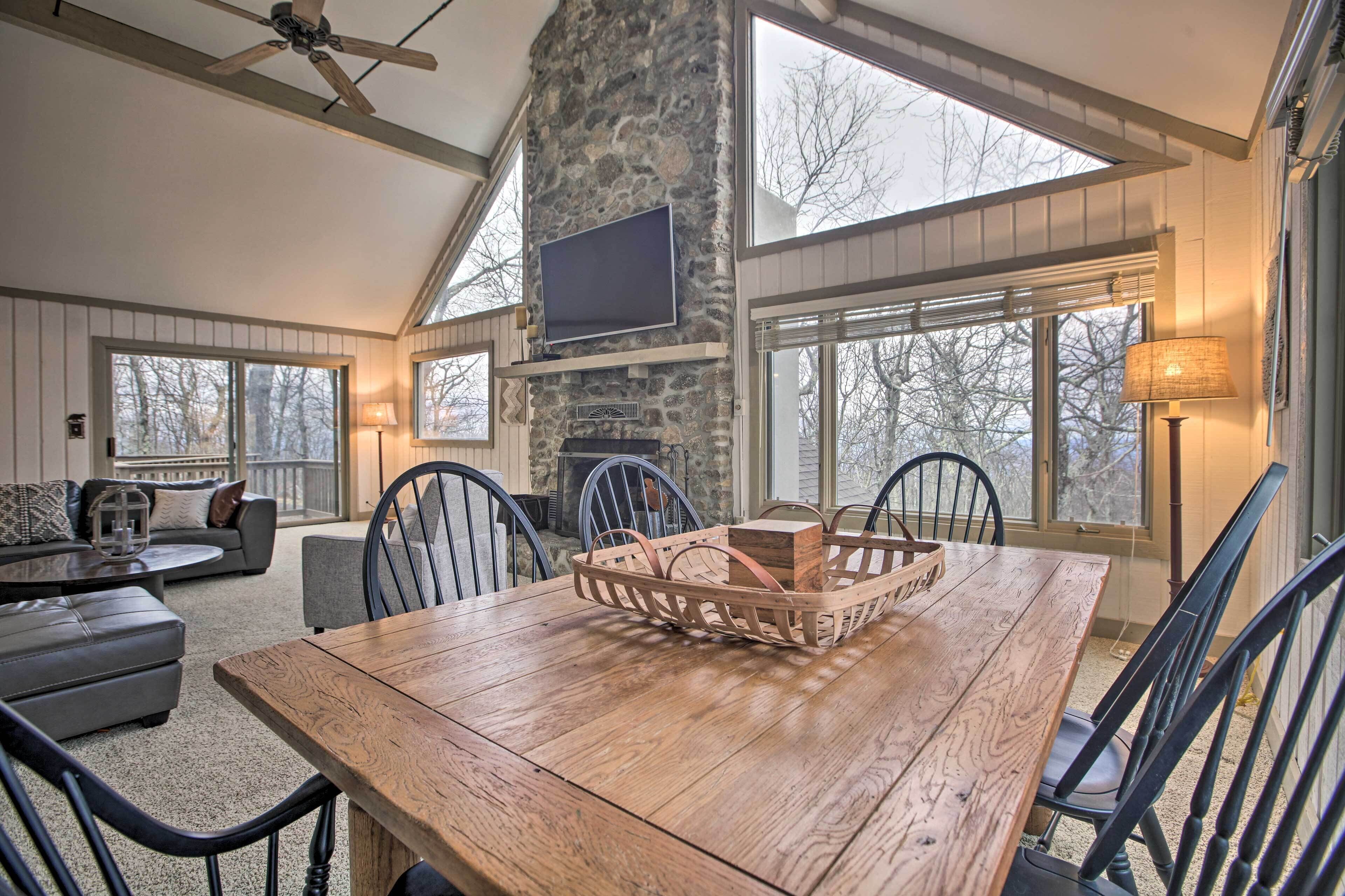 A dining area with a wooden table, chairs, a stone fireplace, and large windows.