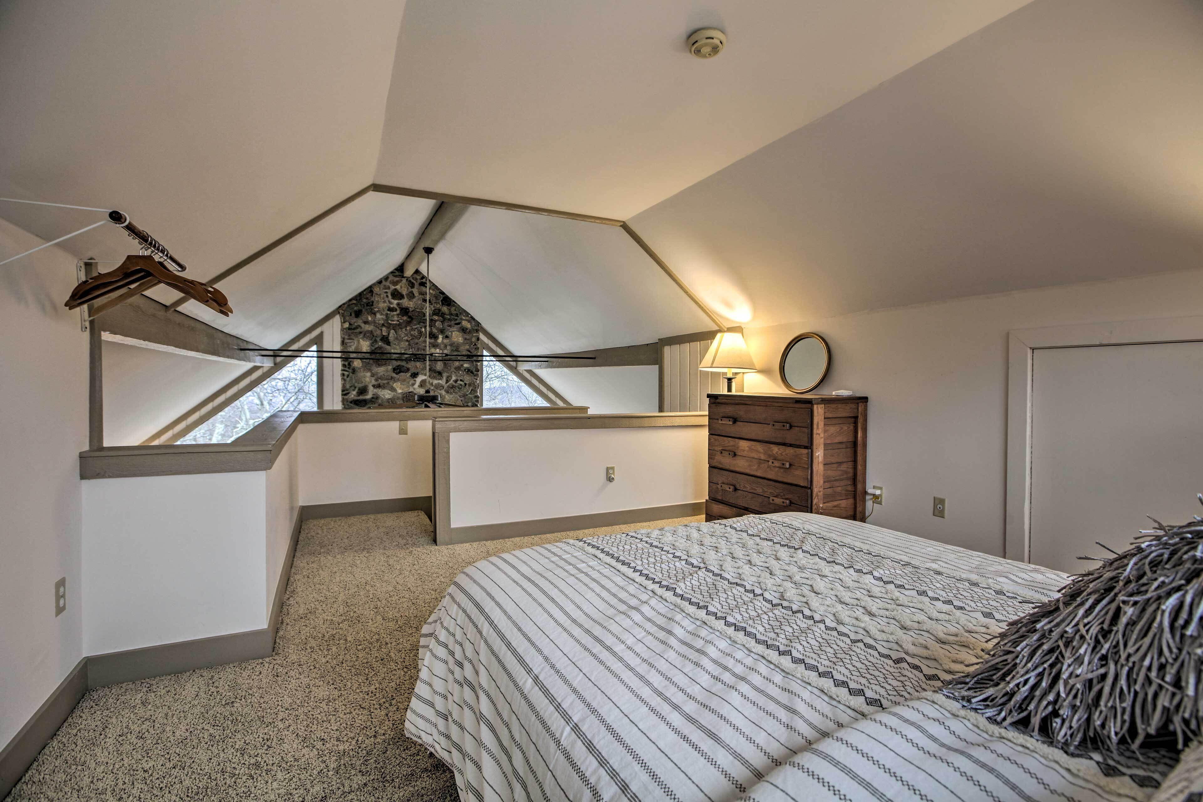 Ein Schlafzimmer mit schrägem Dach, einem Bett mit gemustertem Überwurf, einem Schminktisch, einem Spiegel und einem kleinen Fenster.