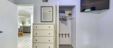 Een kamer met een witte ladekast, een spiegel, een aan de muur bevestigde televisie en een deur naar een andere kamer.