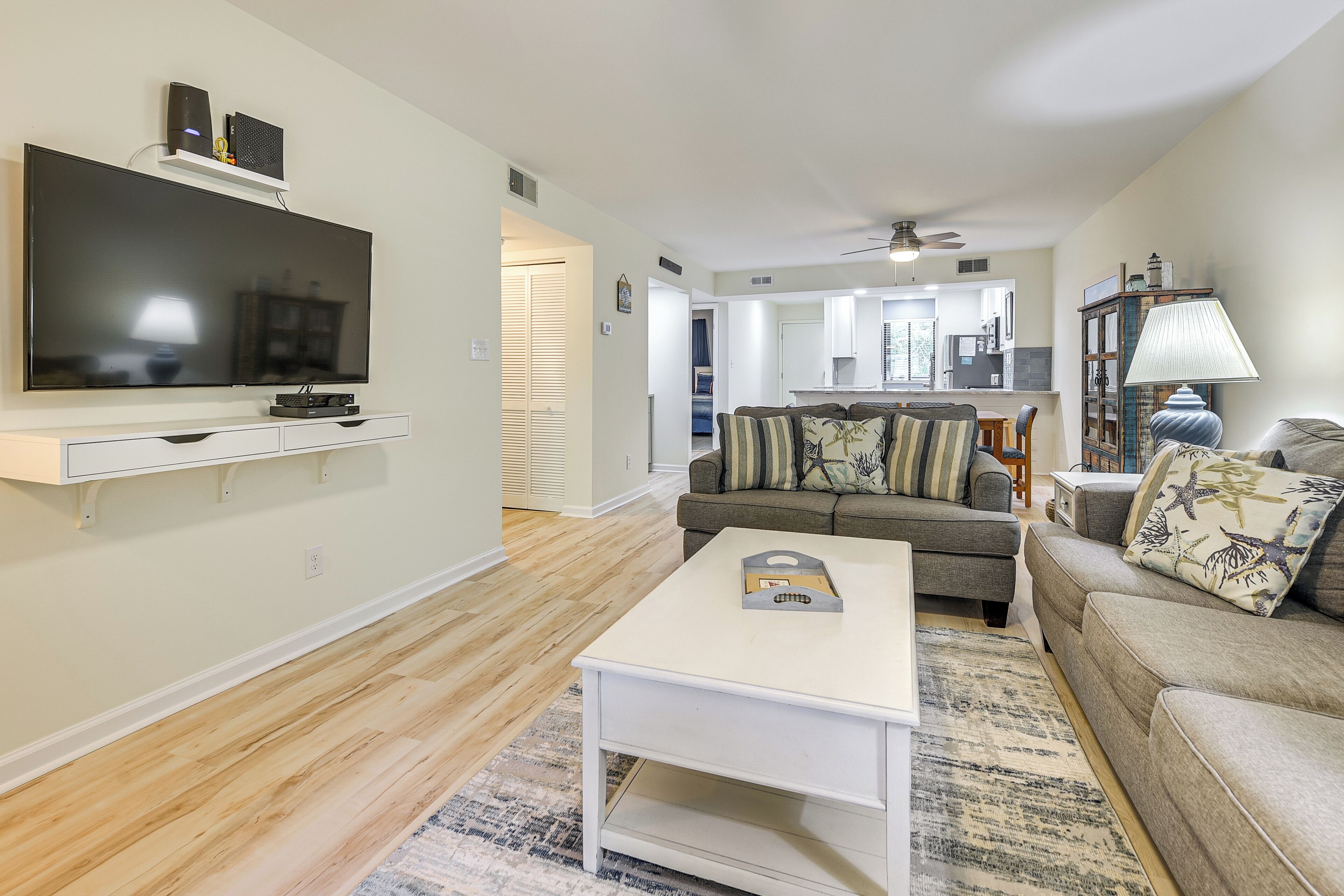 A modern living room with a flat-screen TV, a white coffee table, a grey sofa, and a patterned rug.