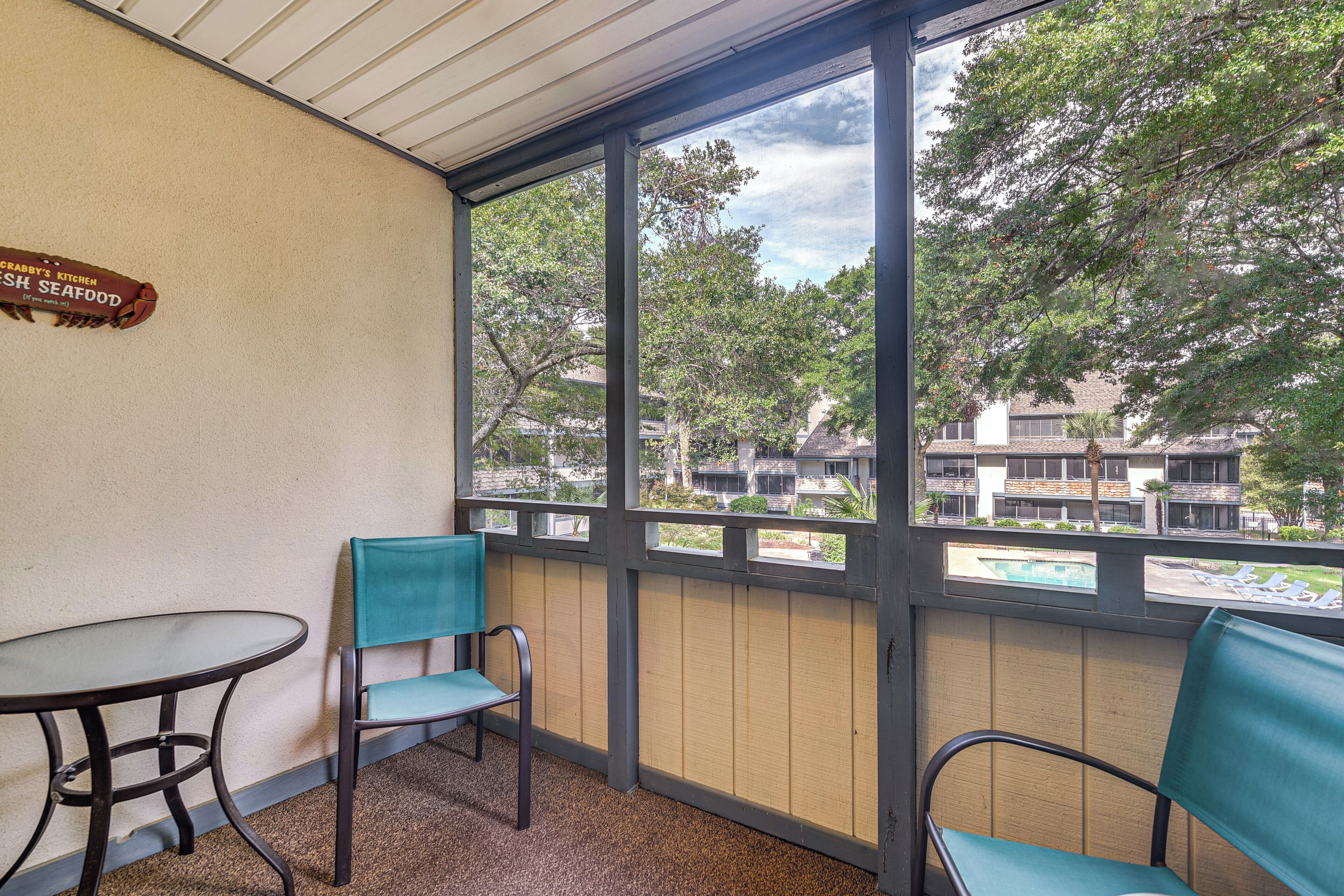 A balcony with a table and two chairs, a sign on the wall, and a view of buildings and trees.