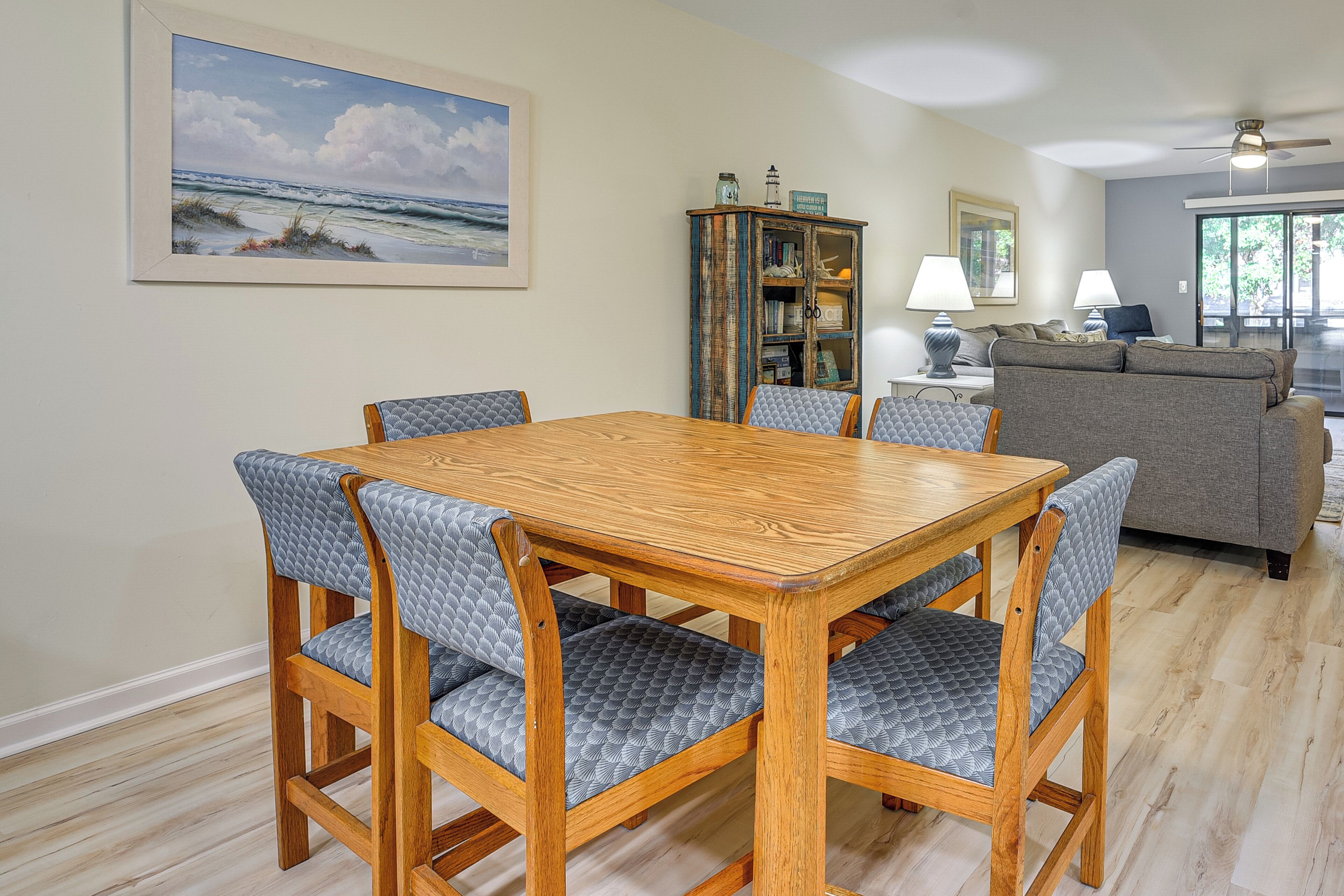 A dining area with a wooden table and cushioned chairs, a bookshelf, a sofa, and a view of the outdoors.