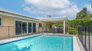 Une piscine avec un patio couvert, une terrasse entourée d’une clôture et un ciel d’un bleu éclatant.