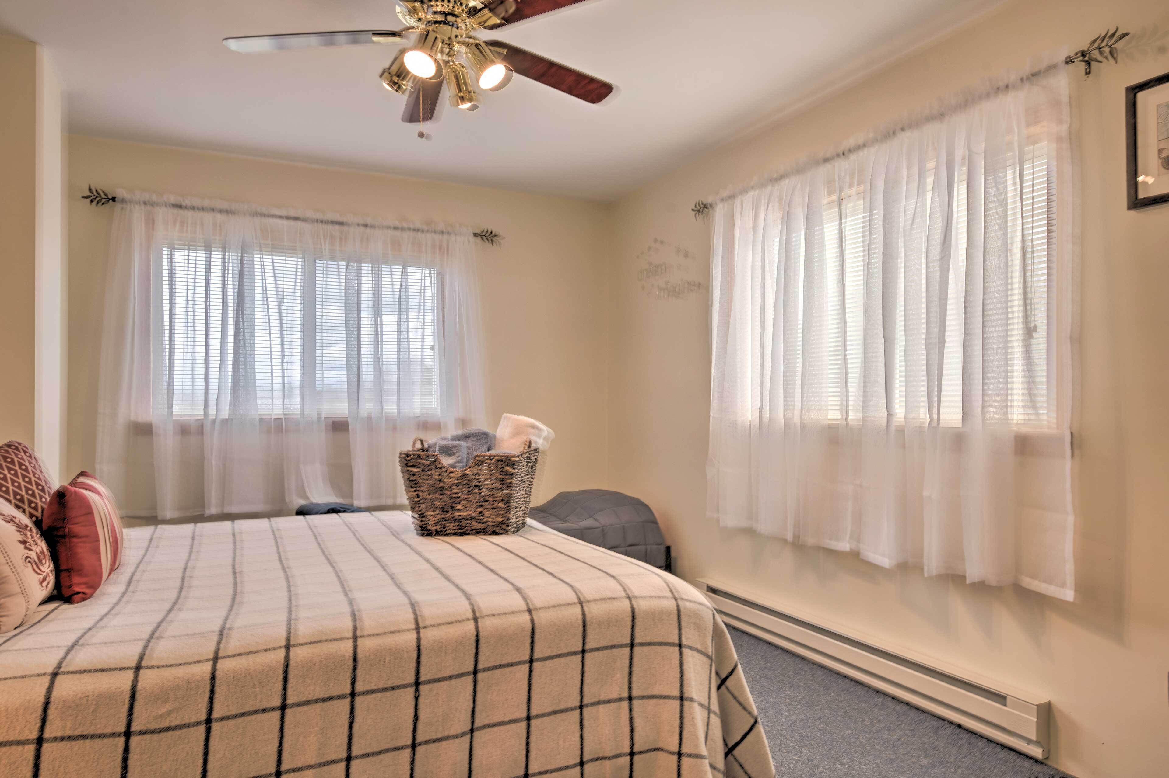 Un dormitorio con cama, ventilador de techo, dos ventanas con cortinas translúcidas, calentador de pared y una cesta con objetos sobre la cama.