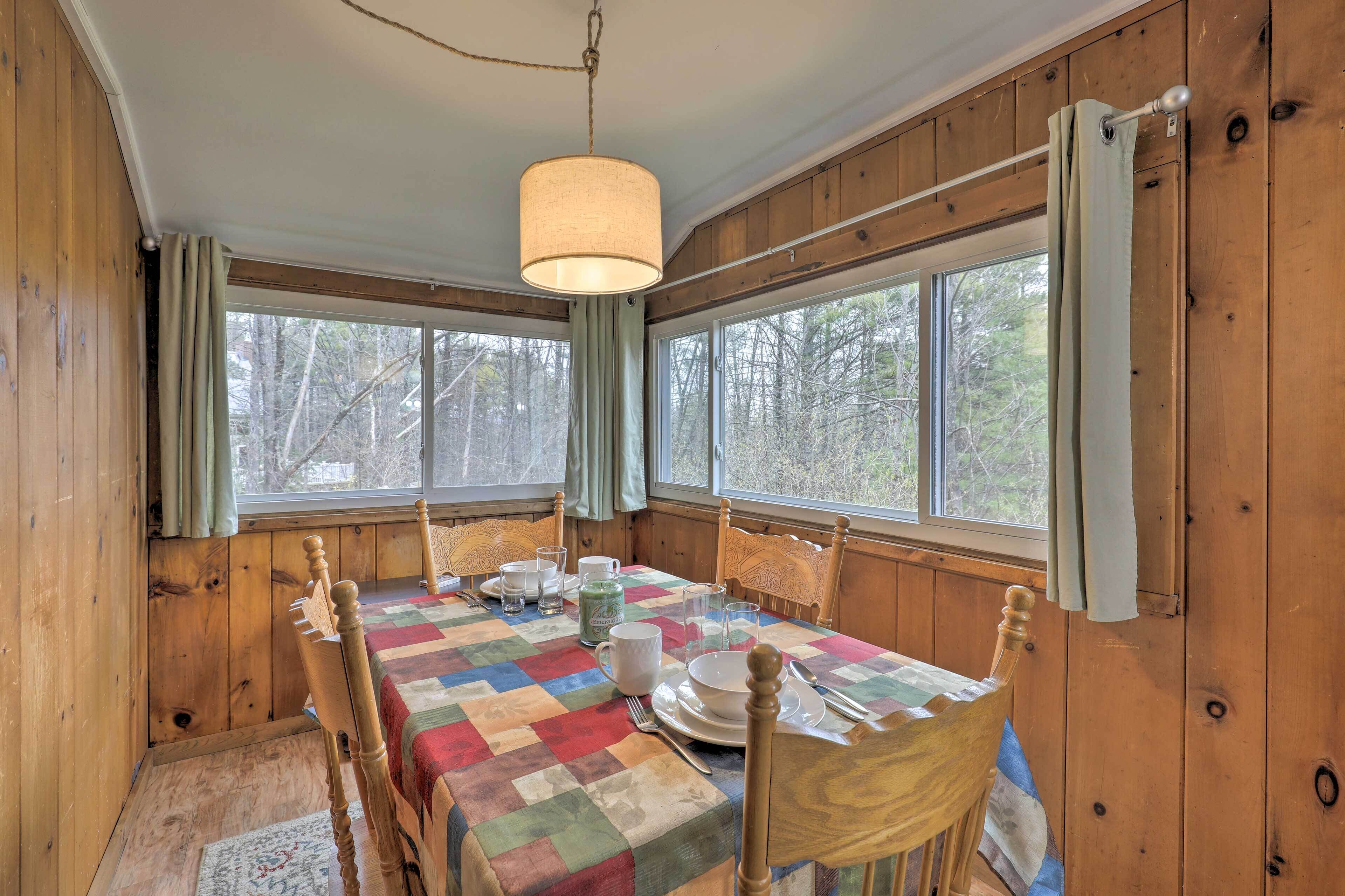 Une salle à manger aménagée dans une cabane en bois, avec une table dressée pour quatre personnes, des chaises en bois, un luminaire suspendu et de grandes fenêtres avec des rideaux.