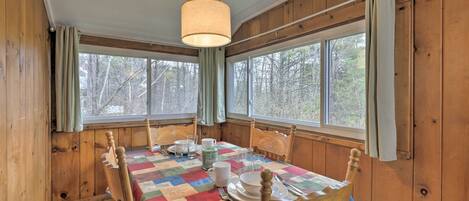Une salle à manger aménagée dans une cabane en bois, avec une table dressée pour quatre personnes, des chaises en bois, un luminaire suspendu et de grandes fenêtres avec des rideaux.
