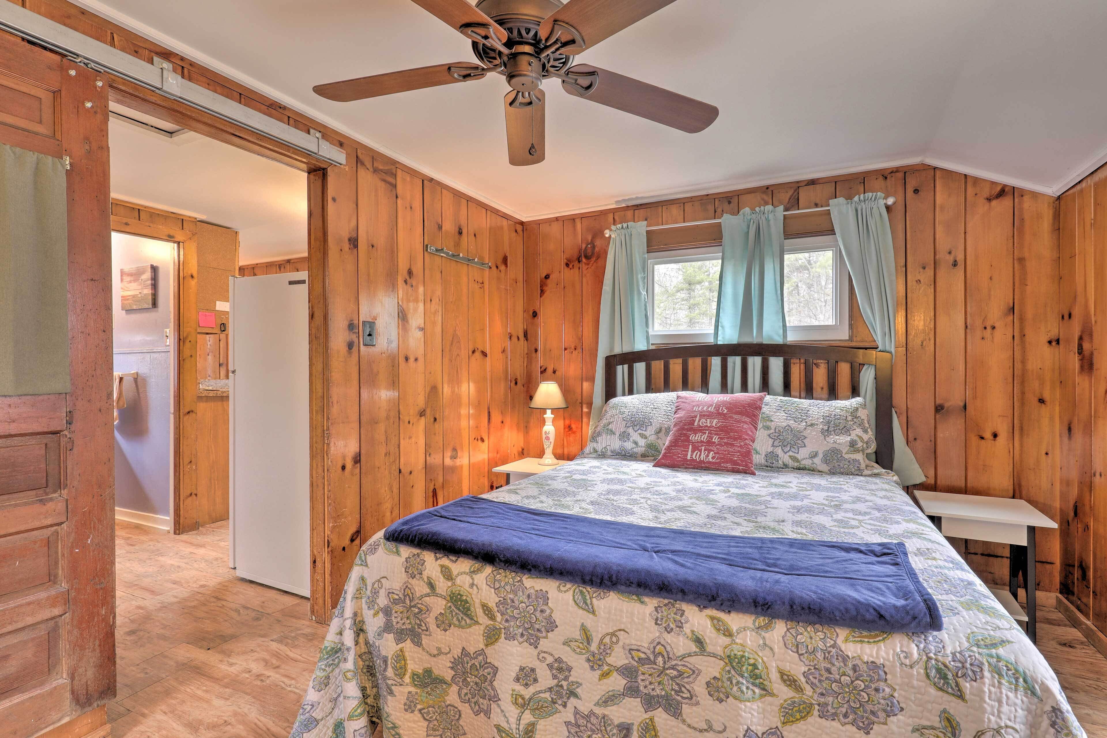 A wooden-paneled bedroom with a bed, a ceiling fan, a window with curtains, and a nightstand with a lamp.