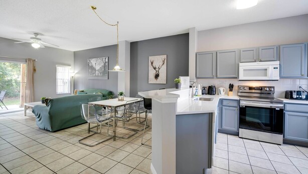 A modern kitchen with grey cabinets, a white island, and stainless steel appliances. There is a dining area with a table and chairs, and a green sofa in the background. - Kissimmee Escape ~ 10 Mi to Disney World! (Kissimmee)