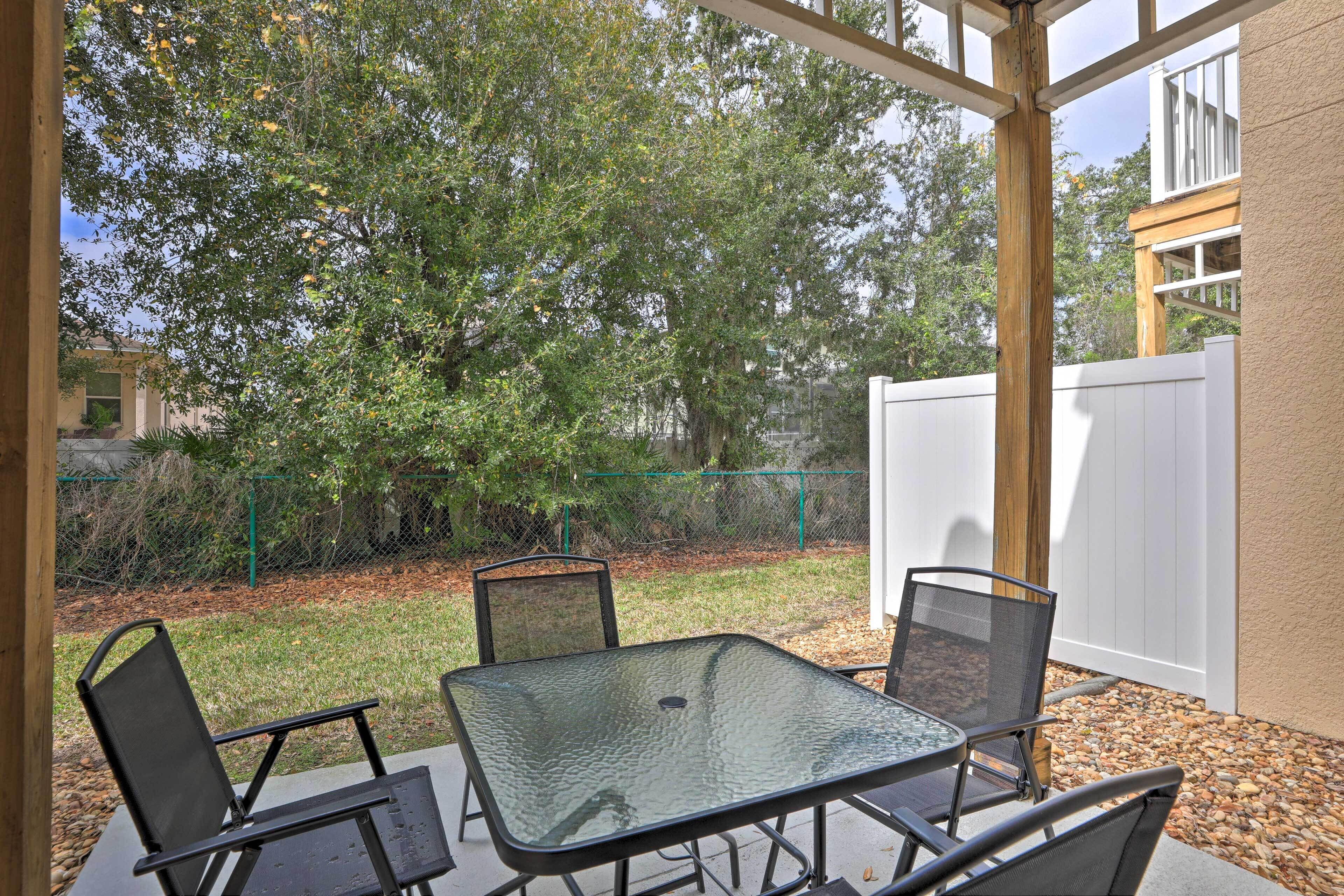 An outdoor patio with a glass table and metal chairs, surrounded by a wooden pergola and a white fence.
