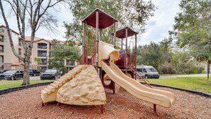 Ein Spielplatz mit Rutsche und Klettergerüst, umgeben von Bäumen und einem Gebäude im Hintergrund.