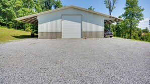 A single-story metal building with a large garage door, surrounded by a gravel driveway and trees. - Wooded Gilbertsville Hideaway on Kentucky Lake! (Gilbertsville)