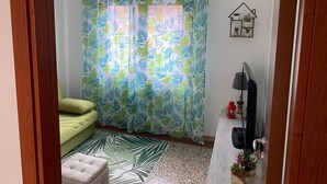 Een woonkamer met een groene bank, een witte fauteuil, een gordijn met een patroon, een klein tafeltje met een rode lantaarn, en een aan de muur bevestigde televisie.