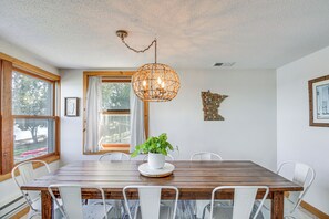 Een eethoek met een houten tafel, witte stoelen, een hanglamp en een potplant.
