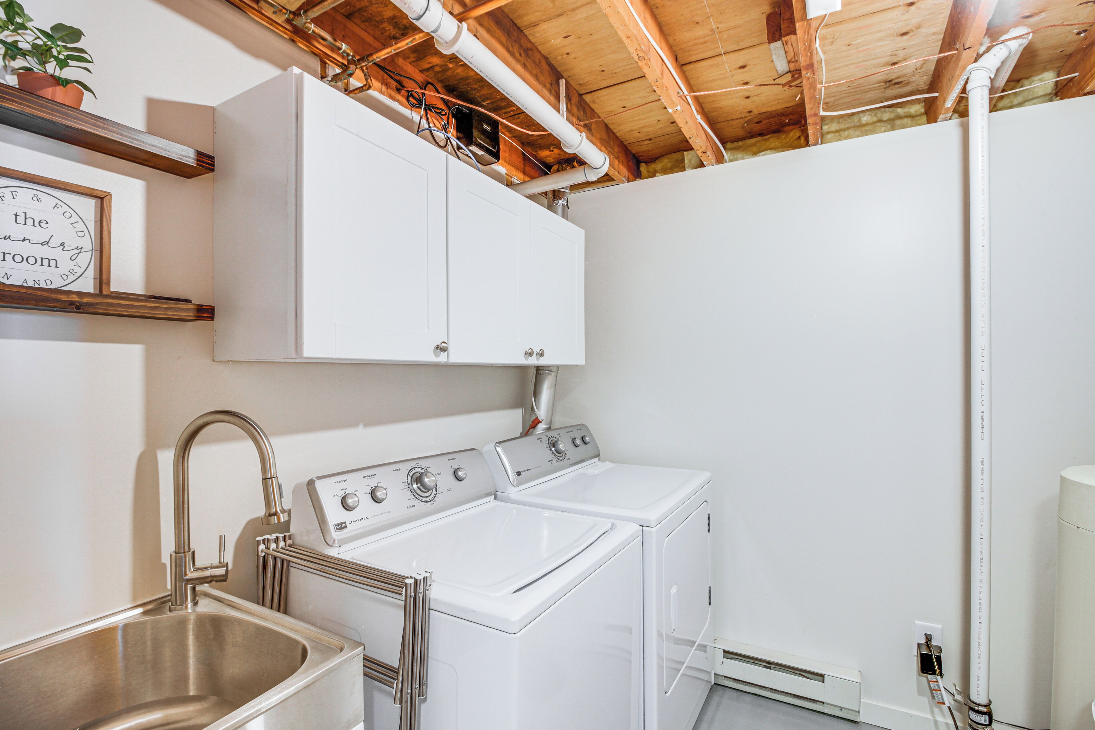 A laundry room with a stainless steel sink, a washing machine, and a dryer. There are wooden beams on the ceiling and a white cabinet above the washing machine.
