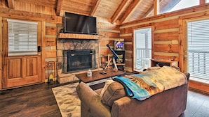A log cabin interior with a stone fireplace, wooden walls, a television mounted above it, and a cozy seating area with a sofa and a coffee table. - Sevierville Cabin w/ Private Hot Tub & Fireplace! (Sevierville)