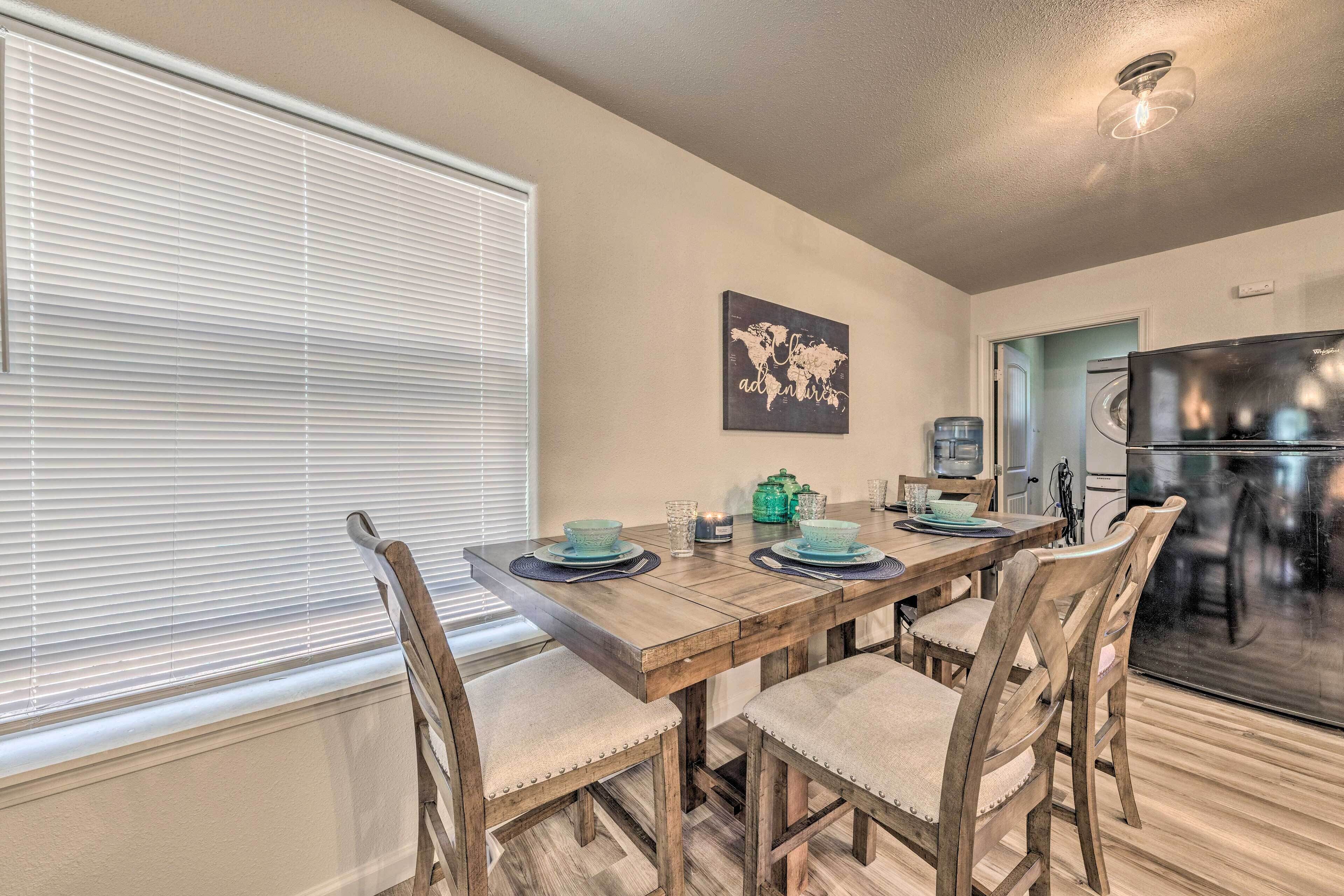 A dining area with a wooden table set for four, surrounded by upholstered chairs. A world map artwork hangs on the wall, and a refrigerator is visible in the background.