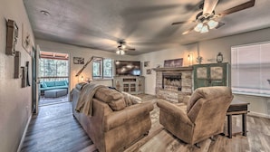 A spacious living room with a fireplace, a ceiling fan, and a view of the outdoors. - Pet-friendly Ruidoso Home w/ Deck & Forest Views! (Ruidoso)