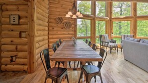 A log cabin interior with a dining table, chairs, a sofa, and large windows offering a view of trees. - Stunning Crosslake Cabin w/ Deck and Lake Views! (Crosslake)