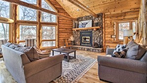 A log cabin interior with a stone fireplace, wooden furniture, and large windows. - Stunning Crosslake Cabin w/ Deck and Lake Views! (Crosslake)