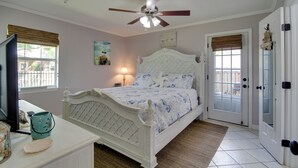 Une chambre à coucher avec un lit blanc, un ventilateur de plafond, un téléviseur posé sur une commode et une fenêtre avec des stores.