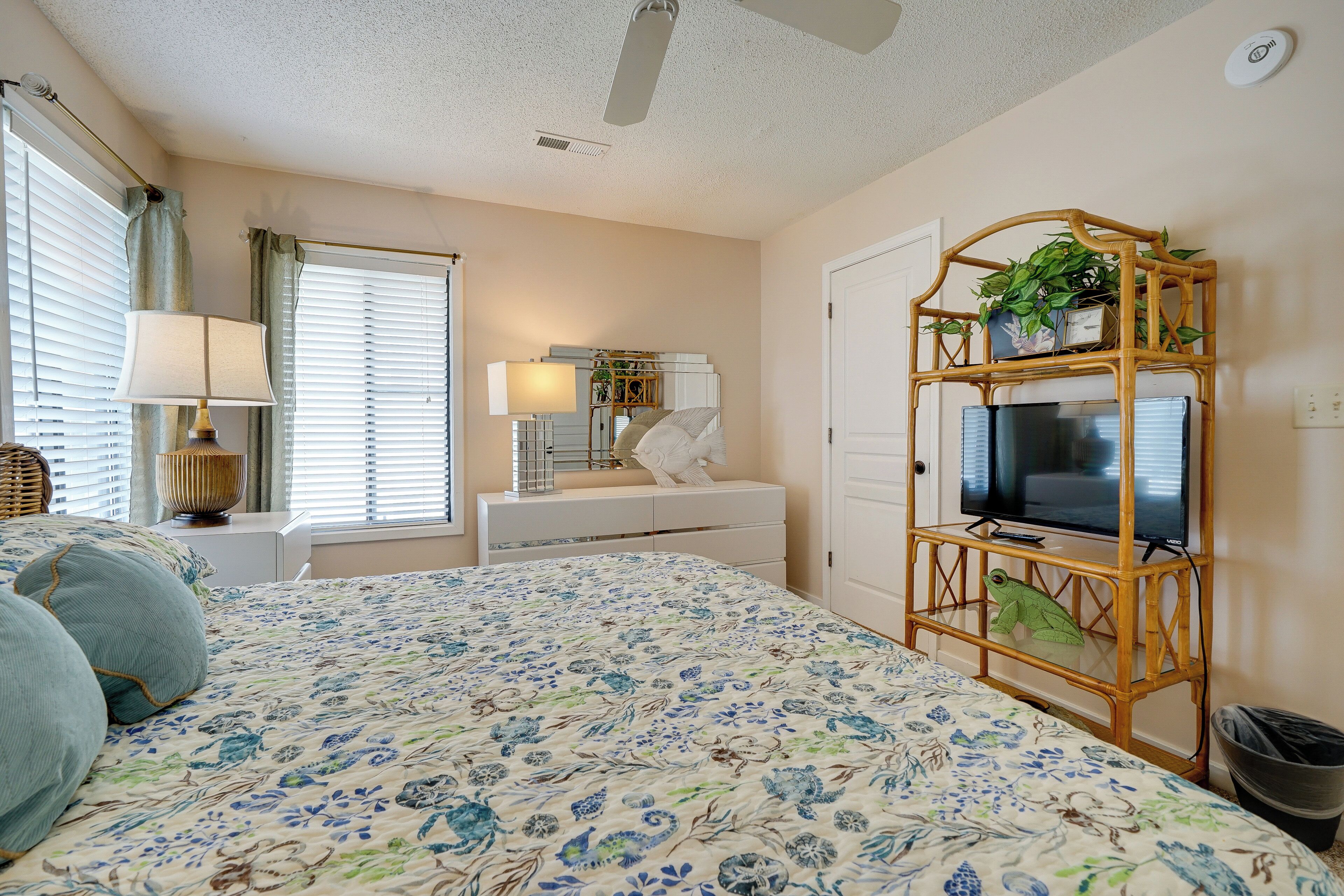 A bedroom with a floral bedspread, a television on a bamboo stand, a dresser, and a window with curtains.
