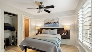 A bedroom with a bed, bedside lamps, a ceiling fan, a painting, and a window with blinds.