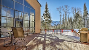 A wooden deck with a glass wall, outdoor chairs, and a view of trees and mountains.
