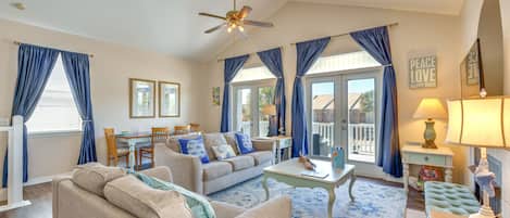 A spacious living room with a beige sofa, a coffee table, and a ceiling fan. There are blue curtains, a dining table with chairs, and a view of the outside through large windows.