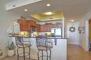 A kitchen with wooden cabinets, a stainless steel refrigerator, and a bar area with two chairs. - Beachfront Bliss on Dauphin Island w/ Pool Access! (Dauphin Island)