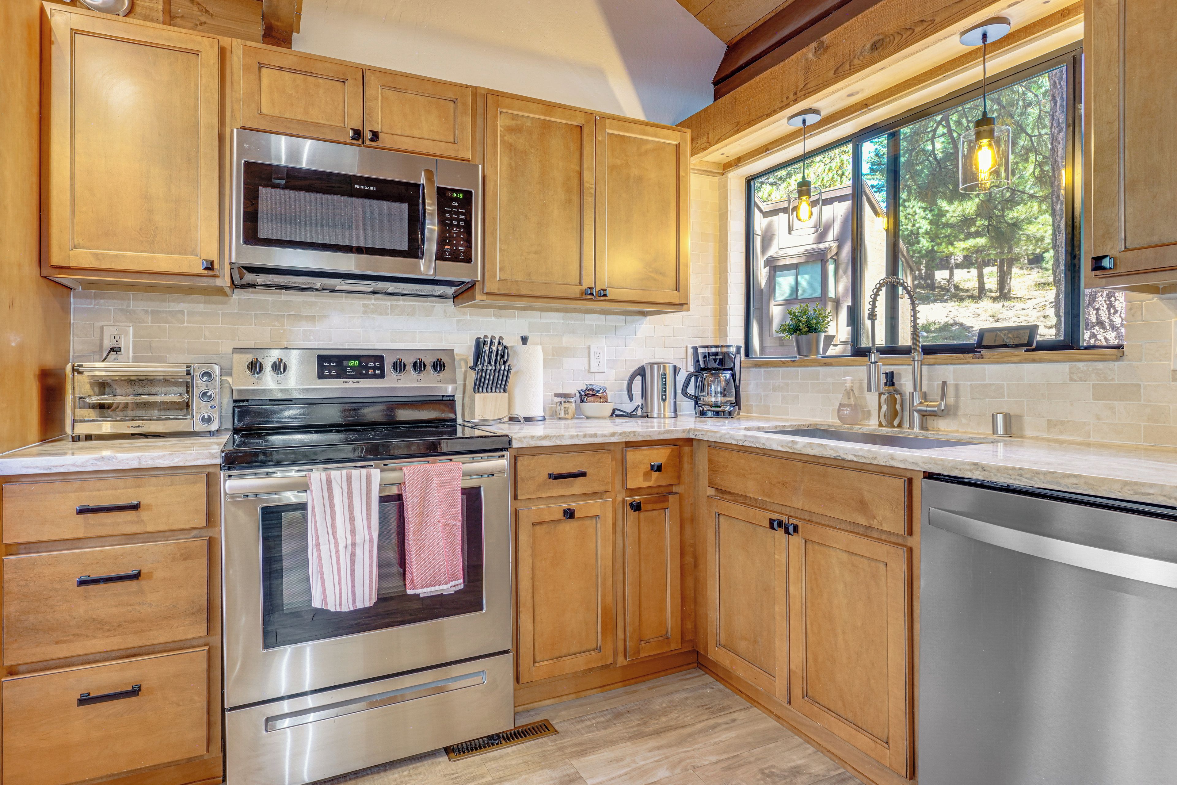 A modern kitchen with wooden cabinets, stainless steel appliances, and a window overlooking a natural landscape.