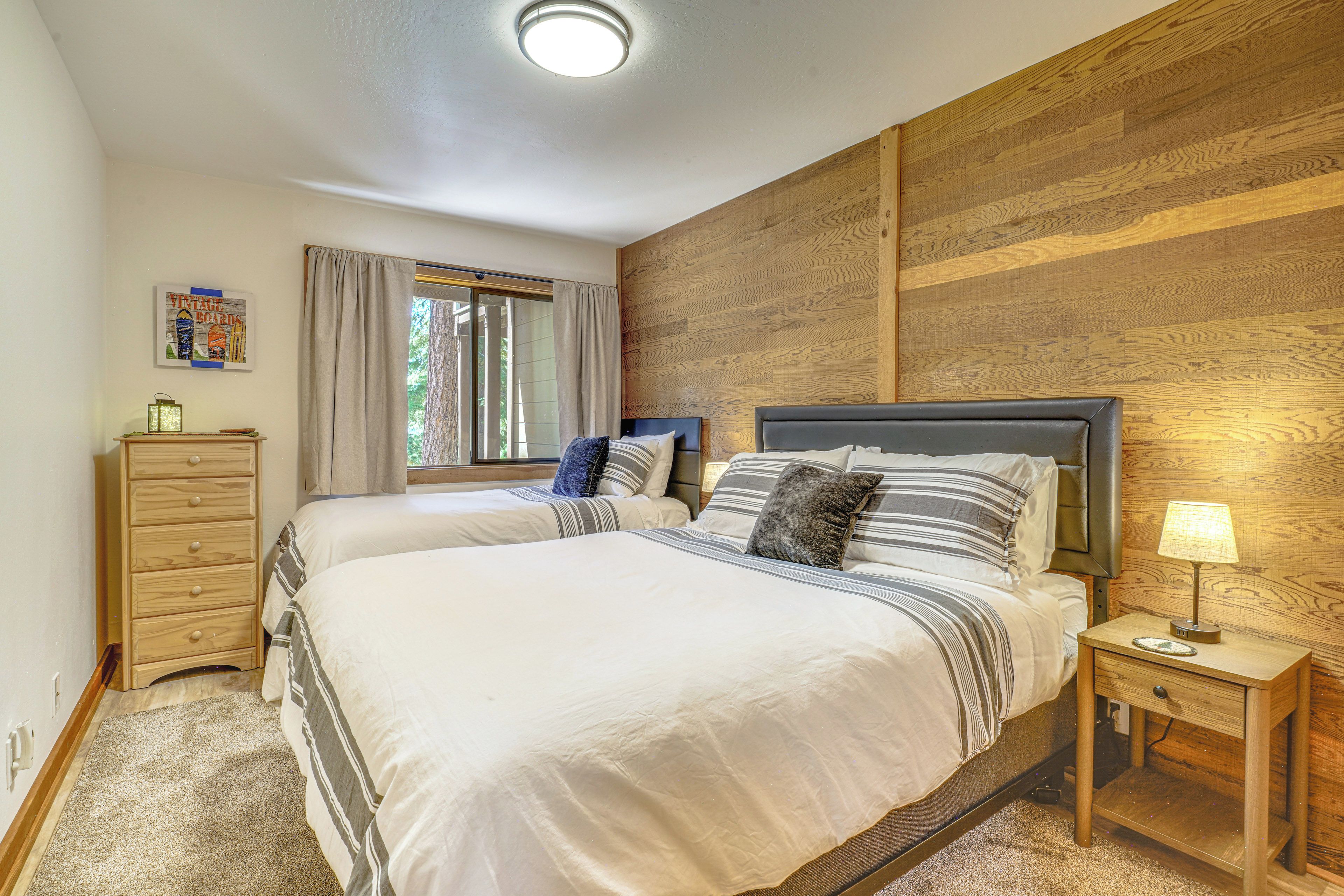 A bedroom with two beds, a wooden headboard, a nightstand with a lamp, a dresser, and a window with curtains.