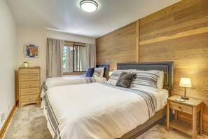 A bedroom with two beds, a wooden headboard, a nightstand with a lamp, a dresser, and a window with curtains.
