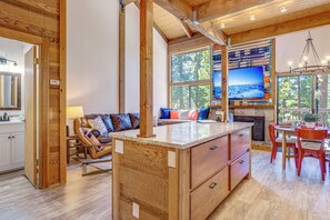 Un'ampia zona giorno con soffitto in legno, un'ampia finestra vista alberi, un'area salotto accogliente con divano e cuscini, e un angolo cucina con tavolo e sedie.