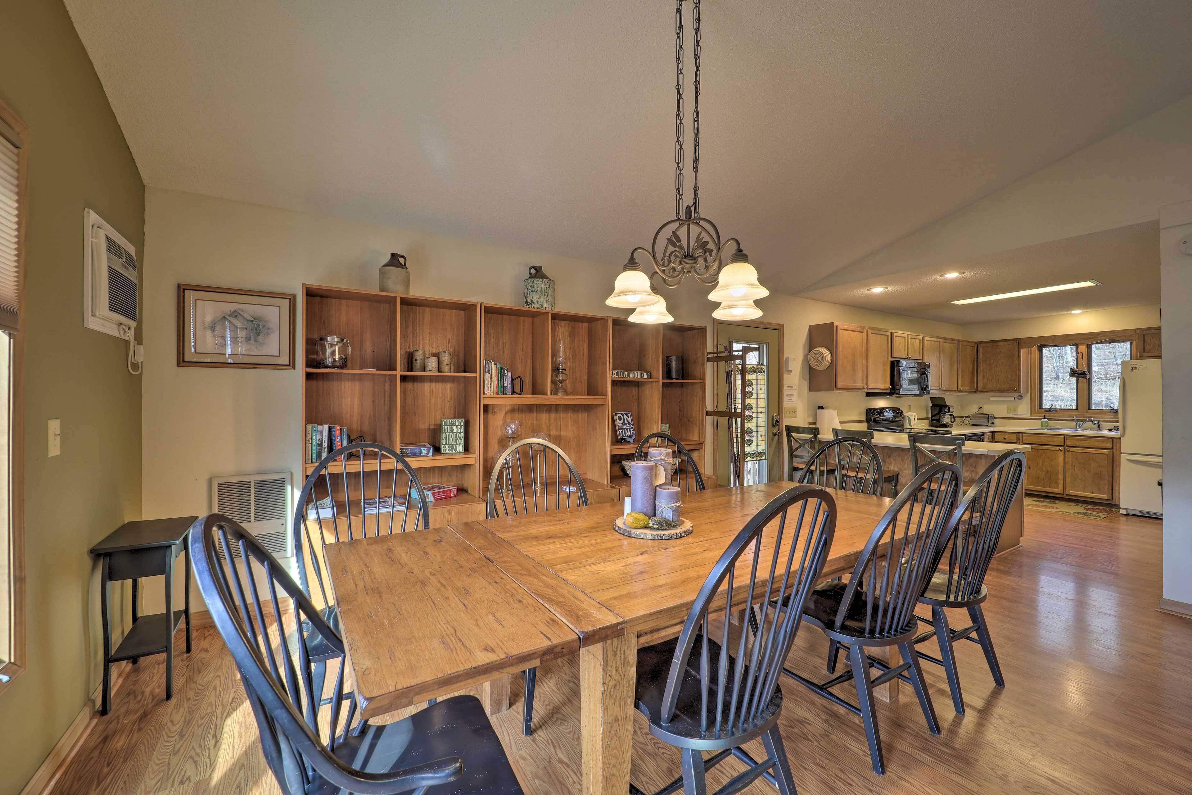 A spacious dining area with a wooden table and chairs, a chandelier, and a kitchen in the background.