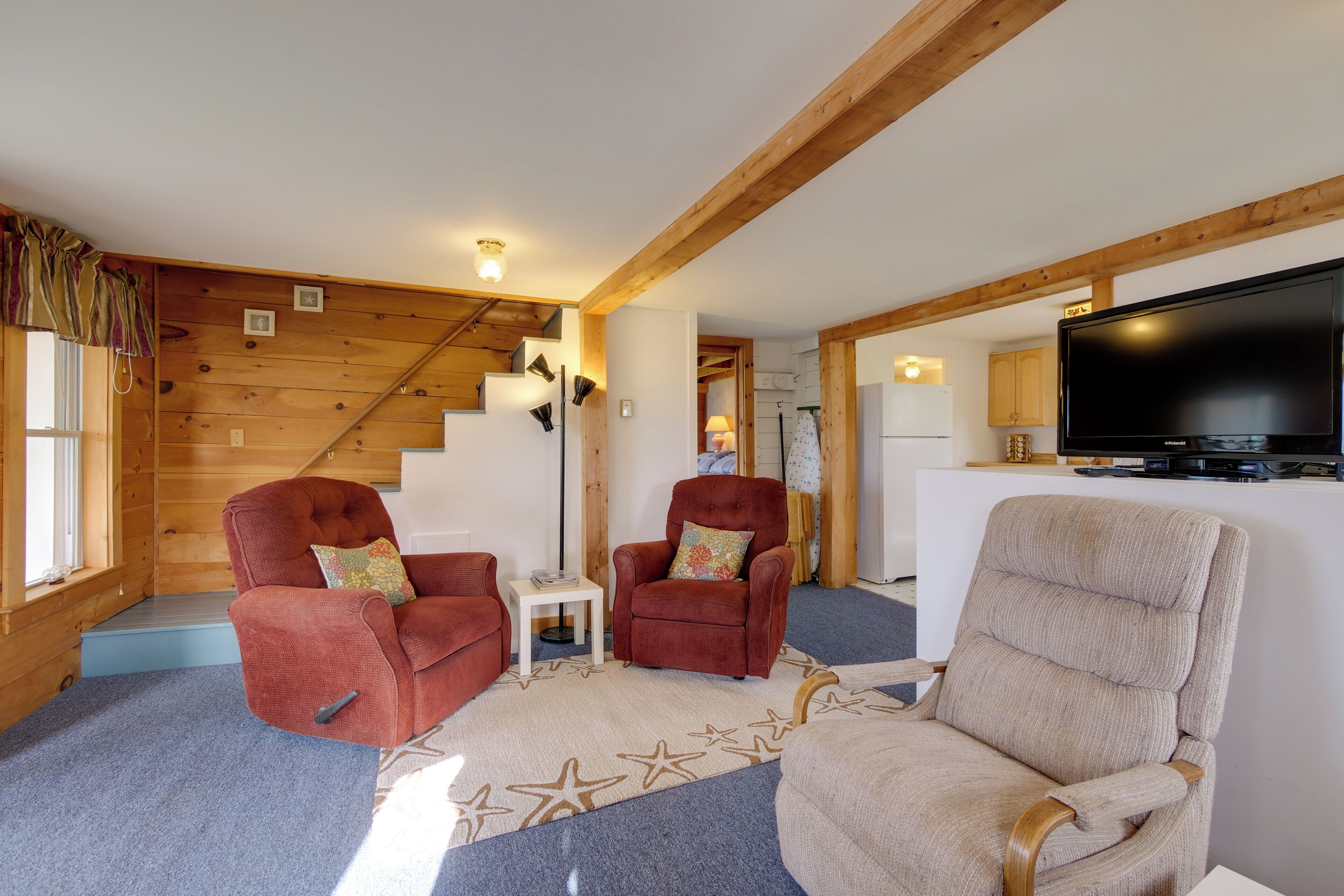 A cozy living room with wooden beams, two red armchairs, a beige armchair, a flat-screen TV, and a staircase leading to another room.