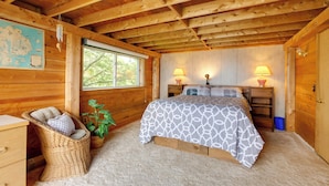 A cozy wooden cabin bedroom with a bed, two bedside lamps, a wicker chair, a small plant, and a window with a view of trees. - Waterfront Cottage - 17 Mi to Acadia Ntnl Pk! (Ellsworth)