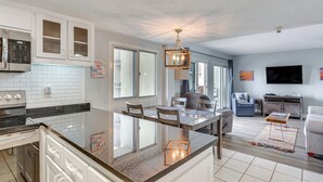 Une cuisine moderne avec un îlot central, un coin repas avec des chaises, un salon avec un canapé et un téléviseur fixé au mur.