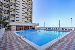 Uma piscina com deck revestido de azulejos, um edifício com varandas e vista para o oceano.