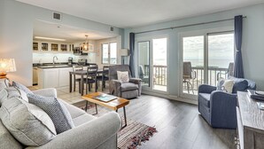 Un salon moderne avec un canapé, un fauteuil et une table à manger avec des chaises. On aperçoit une cuisine au fond, et il y a un balcon avec vue.