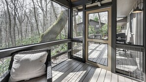 A wooden deck with a glass enclosure, a cushioned bench, and a view of a forested area. - Walk to Main St: Highlands Cottage w/ Fireplace (Highlands)