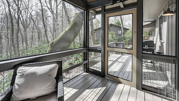 A wooden deck with a glass enclosure, a cushioned bench, and a view of a forested area. - Walk to Main St: Highlands Cottage w/ Fireplace (Highlands)