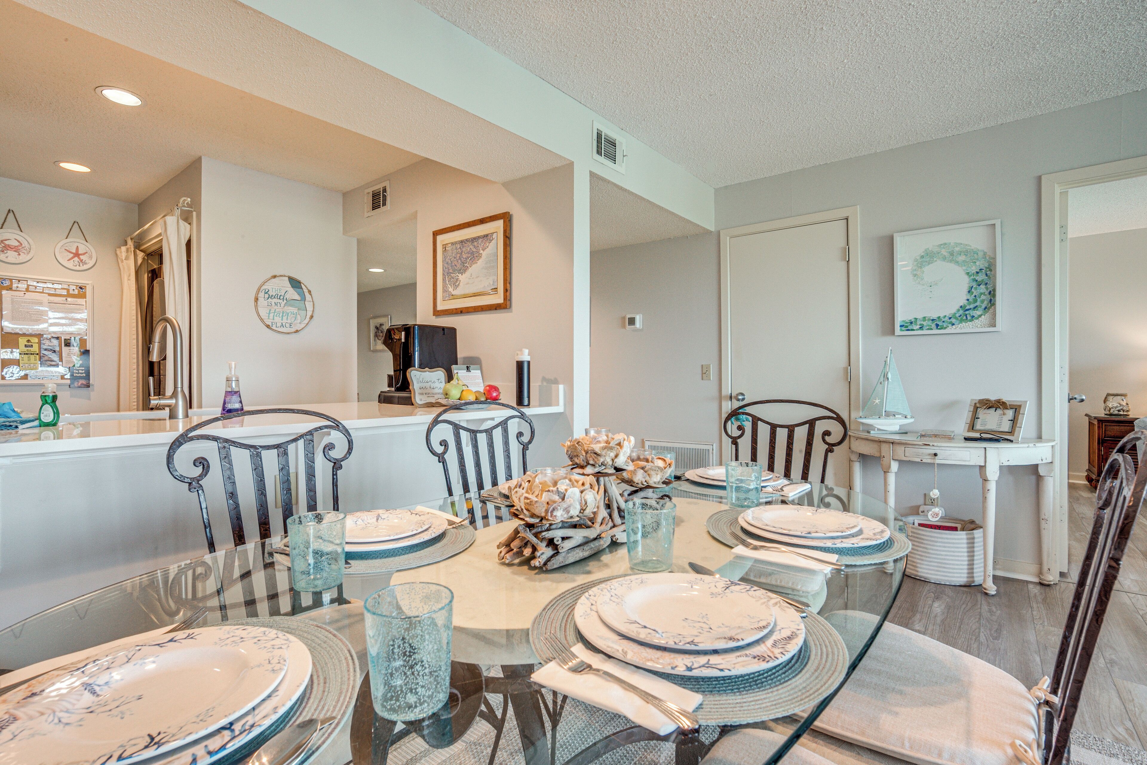 A dining area with a glass table set for six, surrounded by chairs. There is a small side table with a lamp and decorative items.